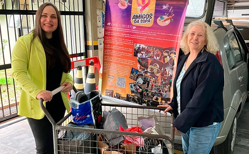 Foto posada de duas mulheres segurando um carrinho carregado de sacolas. Ao fundo está um carro parcialmente coberto por um banner com o texto "Amigos da Sopa" em destaque