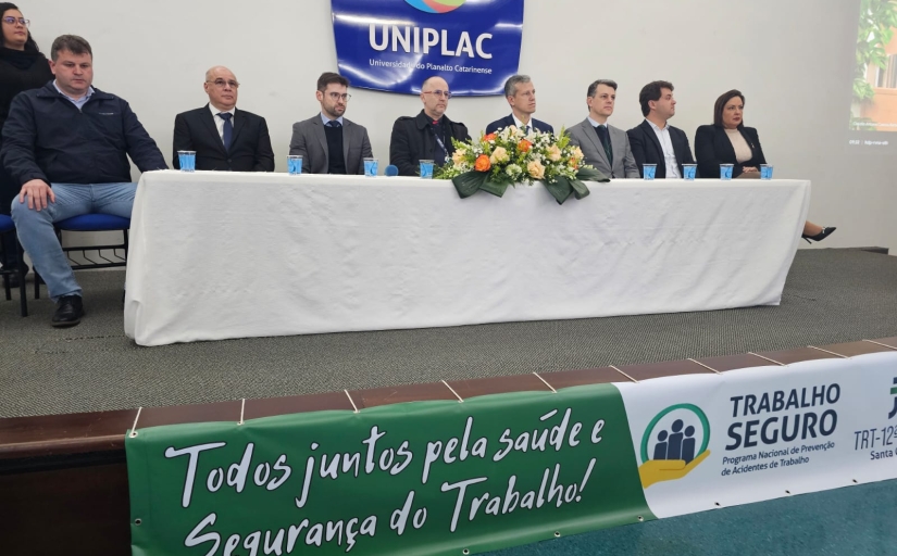 Foto onde estão 8 pessoas sentadas de frente para uma mesa no topo de um palco. A maioria são homens, apenas uma mulher está sentada e outra está em pé no canto esquerdo da foto. Ao fundo está um banner onde está escrito "Uniplac" e no degrau do palco há outro onde se lê "Todos juntos pela saúde e Segurança do Trabalho!"