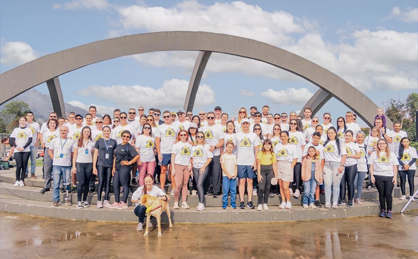 Grupo de cerca de 80 pessoas com camisetas brancas posam para a foto ao ar livre. Como cenário de fundo, um grande arco com três vigas e o céu azul com nuvens