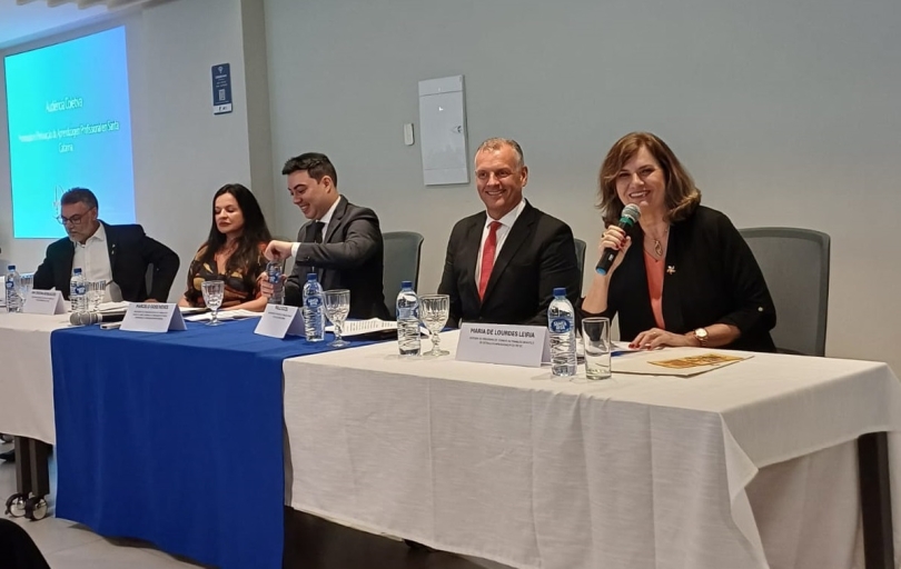 Foto de mesa de evento composta por cinco pessoas, sendo três homens e duas mulheres. Uma delas fala ao microfone