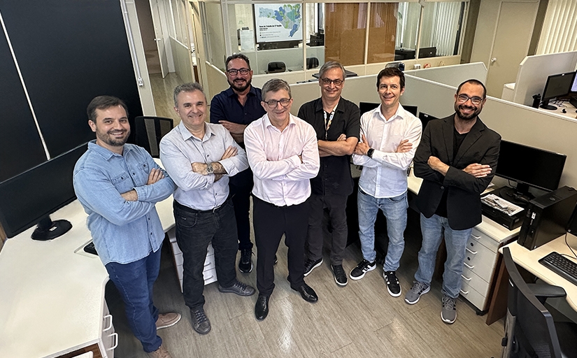 A foto mostra um grupo de sete homens em pé, sorrindo e de braços cruzados, posando para a câmera em um ambiente de escritório. Todos estão em um local bem iluminado, com divisórias e várias mesas com computadores ao fundo.