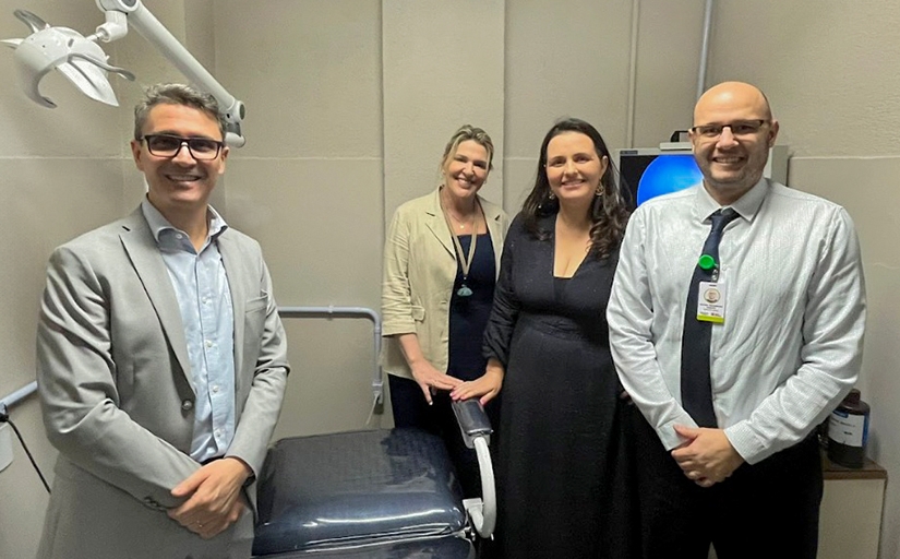 Foto posada de quatro pessoas em uma sala com alguns equipamentos eletrônicos ao fundo e uma cadeira reclinável de consultório médico