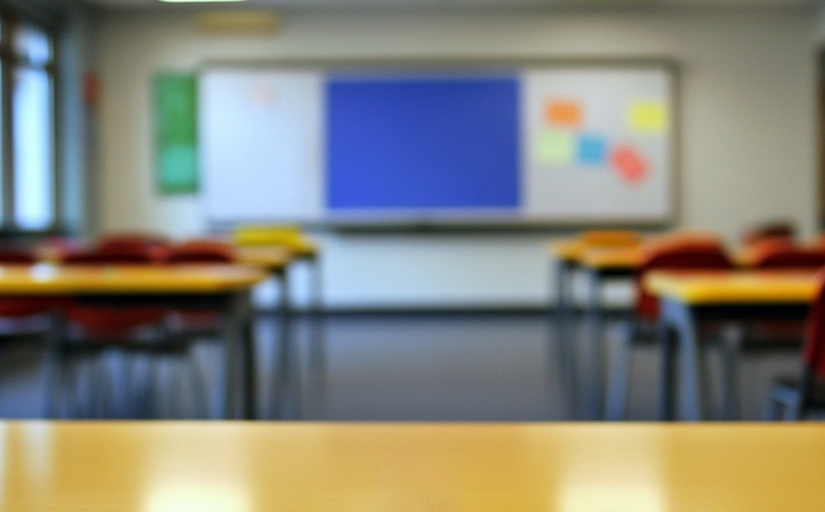 Arte de uma sala de aulas a partir da perspectiva da mesa do professor. É possível perceber, desfocado, cadeiras vazias um quadro de avisos ao fundo.