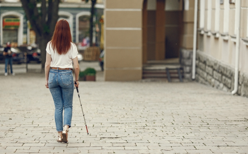 Foto mostra uma mulher cega de costas caminhando pela rua, com uma bengala