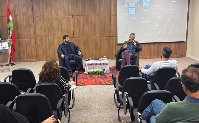 Fotografia de um auditório, no qual há dois homens sentados em duas poltronas individuais à frente. Um deles fala ao microfone para a plateia, que está sentada em cadeiras pretas. Ao fundo, sobre um painel de madeira, há uma tela de projeção à direita. À esquerda, há uma bandeira do estado de Santa Catarina e um púlpito.