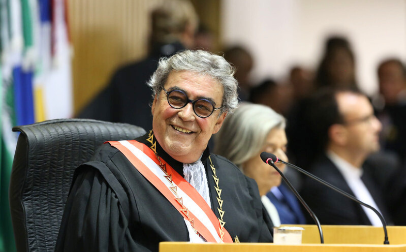 A foto mostra uma pessoa com cerca de 60 anos, cabelos grisalhos, sorrindo, e vestindo uma toga judicial. Ela está sentada em um ambiente de tribunal.