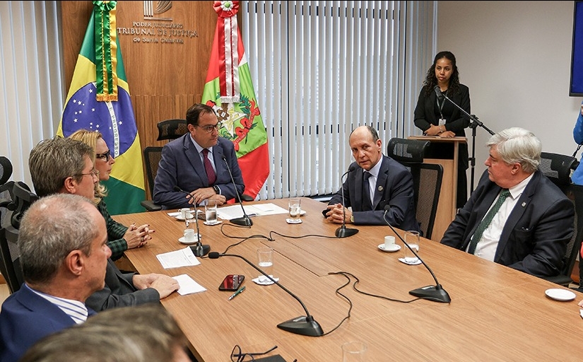 Em uma mesa de madeira, estão sentadas sete pessoas, sendo seis homens e uma mulher. Na mesa, o presidente do TRT-SC, Amarildo Carlos de Lima, um homem branco, de olhos claros, trajando terno e gravata, fala ao microfone. Ao fundo, há um painel de madeira com o brasão do Tribunal de Justiça de Santa Catarina. À direita, ao fundo, uma mulher negra, de cabelos cacheados e longos, está em pé, em um púlpito.