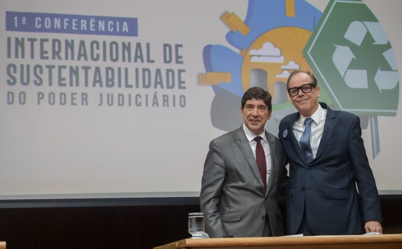 A imagem mostra dois homens de terno posando para uma foto, sorrindo e de frente para a câmera. Eles estão em pé, lado a lado, em frente a um painel que apresenta o título "1ª Conferência Internacional de Sustentabilidade do Poder Judiciário". No fundo, há ilustrações coloridas, incluindo um símbolo de reciclagem e imagens que remetem à natureza e sustentabilidade.