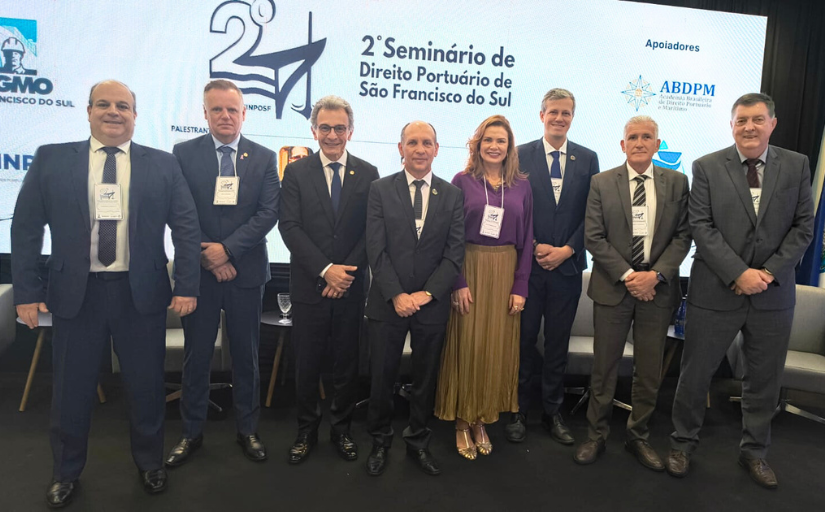 A imagem mostra um grupo de sete homens e uma mulher em um palco, em frente a uma tela de apresentação. A tela exibe o texto “2º Seminário de Direito Portuário de São Francisco do Sul”. As pessoas estão vestidas formalmente, os homens de terno, e a mulher de saia e blusa