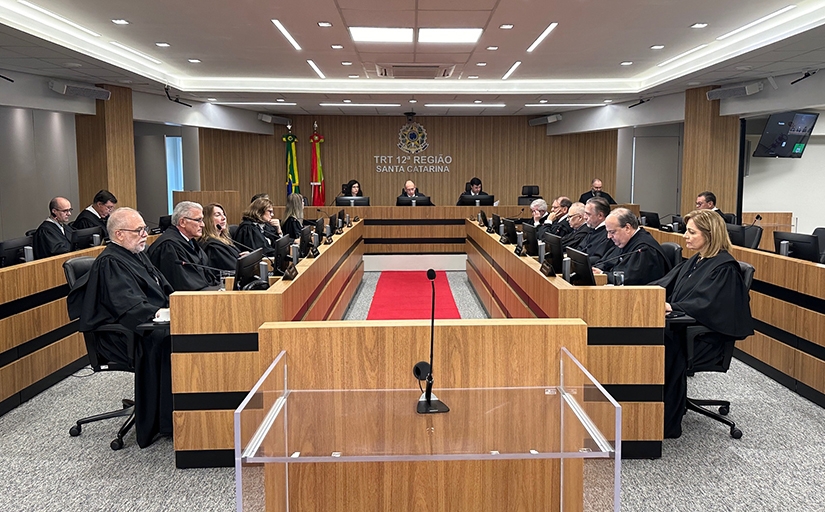 Fotografia da sala de sessões do Tribunal Pleno, com mesa em "U", juízes em togas e bandeiras ao fundo