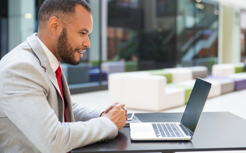 Fotografia ilustrativa de homem negro sorridente vestido formalmente olhando para tela de um notebook ao ar livre