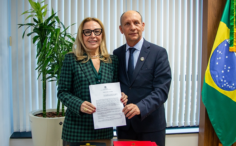 A imagem retrata duas pessoas em pé, lado a lado, segurando um documento. À esquerda, uma mulher vestida com um blazer xadrez verde e preto, óculos de armação grossa e cabelo loiro solto, sorri para a câmera. À direita, um homem de terno escuro, gravata e com expressão mais séria, também olha diretamente para a câmera. Ao fundo, há uma bandeira do Brasil, uma planta em vaso e uma parede com persianas