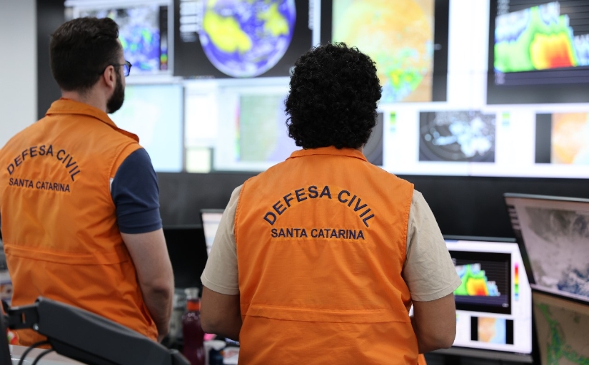 Fotografia de dois agentes da Defesa Civil monitorando telas de clima. Ambos estão de costas para a câmera.