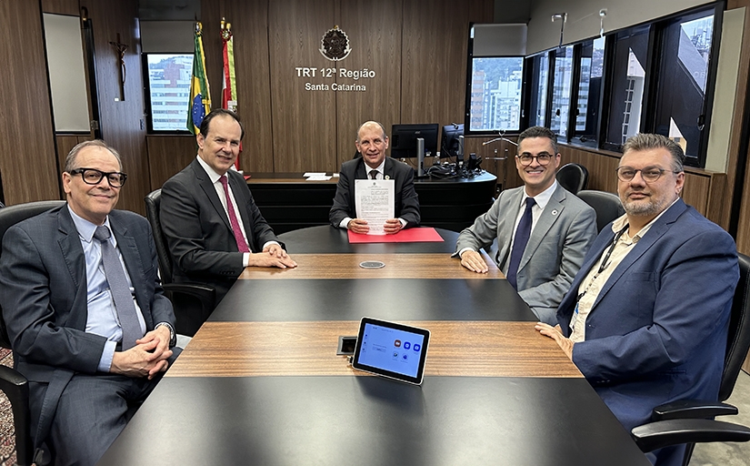 Fotografia com cinco homens de terno em reunião no TRT-SC, com bandeiras ao fundo, documento assinado e um tablet sobre a mesa