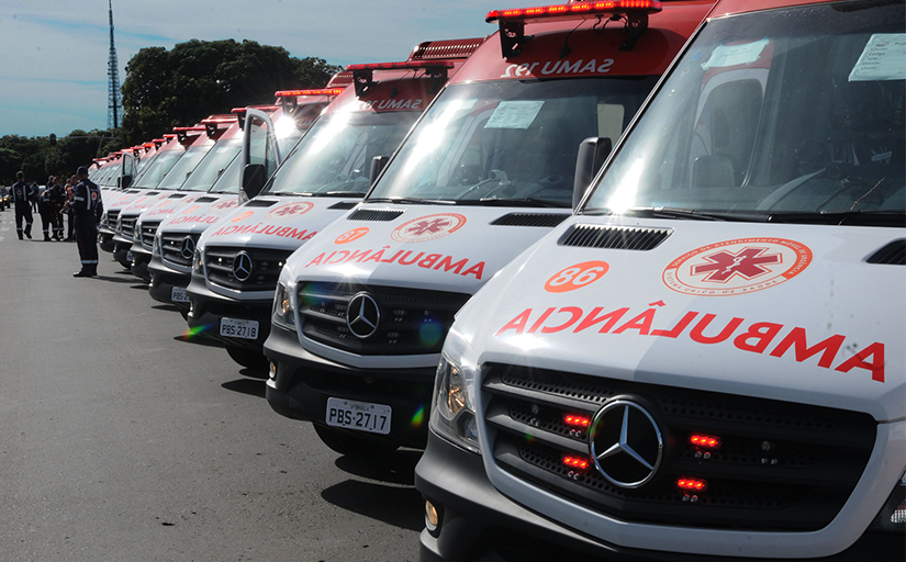 Fotografia de várias ambulâncias do Samu enfileiradas, à direita, em um pátio externo. À esquerda, há um grupo de funcionários do serviço de emergência uniformizados.