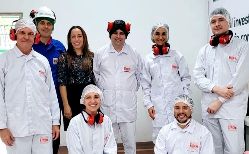 A foto mostra um grupo de cinco homens e três mulheres, sorridentes, a maioria vestindo uniformes brancos com o logotipo "Tirol" e usando equipamentos de proteção, como redes para cabelo e protetores auriculares vermelhos, típicos de um ambiente de produção ou fábrica. 