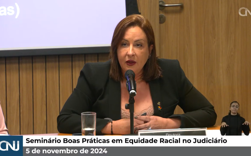 Foto de uma mulher branca de cabelo castanho até os ombros falando ao microfone. Ela veste um blazer preto e uma peça bege coberta pelas mãos. Em uma barra abaixo está escrito "Seminário Boas Práticas em Equidade Racial no Judiciário", ao lado o logo do CNJ e abaixo "5 de novembro de 2024"