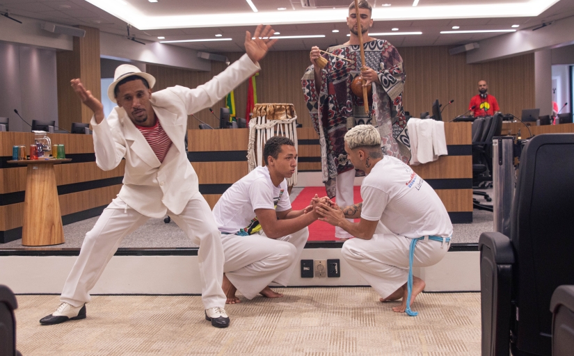 A imagem mostra uma apresentação cultural em um auditório. Em destaque, um homem vestido com um traje branco e chapéu executa uma expressão corporal estilizada, enquanto dois outros, com uniformes brancos de capoeira, estão agachados no centro realizando um cumprimento típico dessa arte. Ao fundo, uma pessoa toca berimbau, e há outros elementos tradicionais, como tambores, além da decoração do auditório com madeira e bandeiras