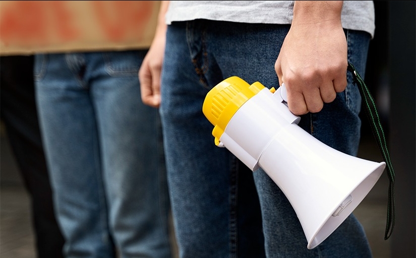 Foto em detalhe com uma pessoa em pé, segurando um megafone abaixado ao lado da perna esquerda.