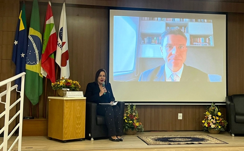 A imagem mostra um palco onde uma mulher está sentada à esquerda, de frente para uma grande tela que exibe outra pessoa. A pessoa sentada está segurando um tablet. Na tela, um homem é mostrado da cintura para cima, em uma videochamada. O fundo da sala inclui várias bandeiras e arranjos florais em ambos os lados da área do palco. 