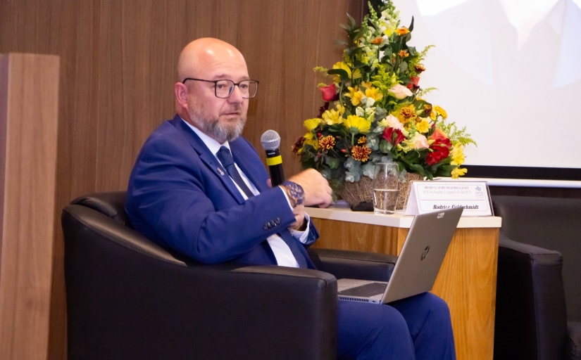 Homem de terno azul usando óculos sentado em poltrona, com microfone na mão e notebook no colo