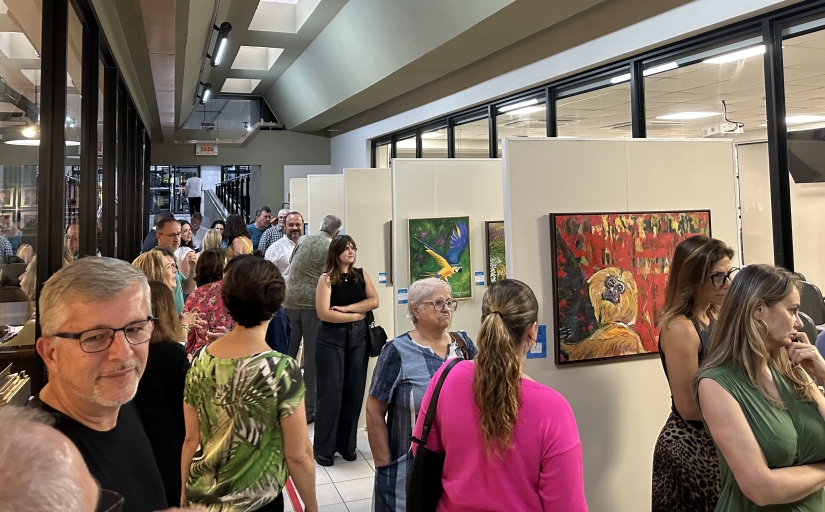 Exposição de arte com quadros coloridos de animais e paisagens, em ambiente moderno, onde um público diverso aprecia as obras.