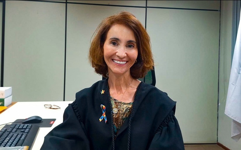 Fotografia da juíza Lisiane Vieira, uma mulher branca, de cabelos curtos na altura dos ombros, ruivos e olhos castanhos. Ela está sentada em sua mesa de trabalho, trajando vestes de magistrada e sorri. 