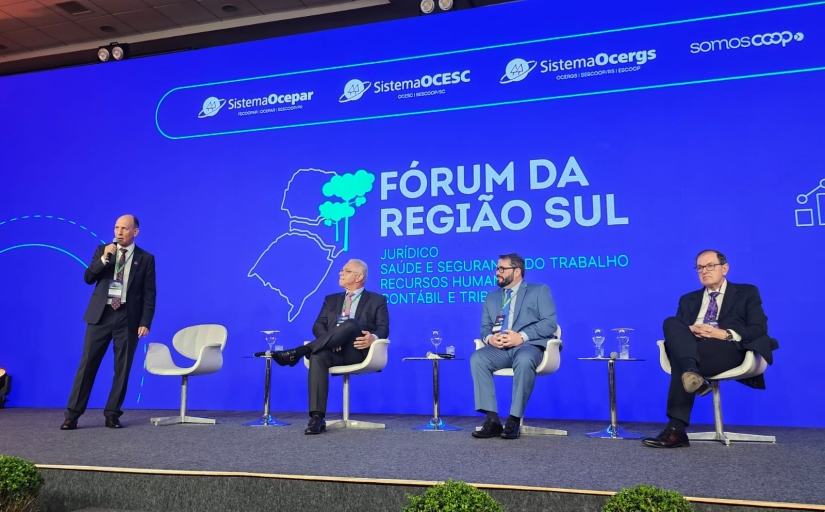 A imagem mostra um palco com quatro cadeiras, três das quais estão ocupadas por homens de terno e gravata. O fundo é uma grande tela azul com o texto “FÓRUM DA REGIÃO SUL” e logotipos de várias organizações