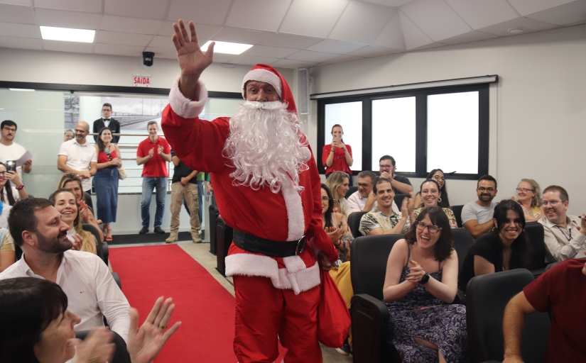 Homem vestido de Papai Noel desce uma rampa dentro de um auditório, sendo aplaudido pelo público presente