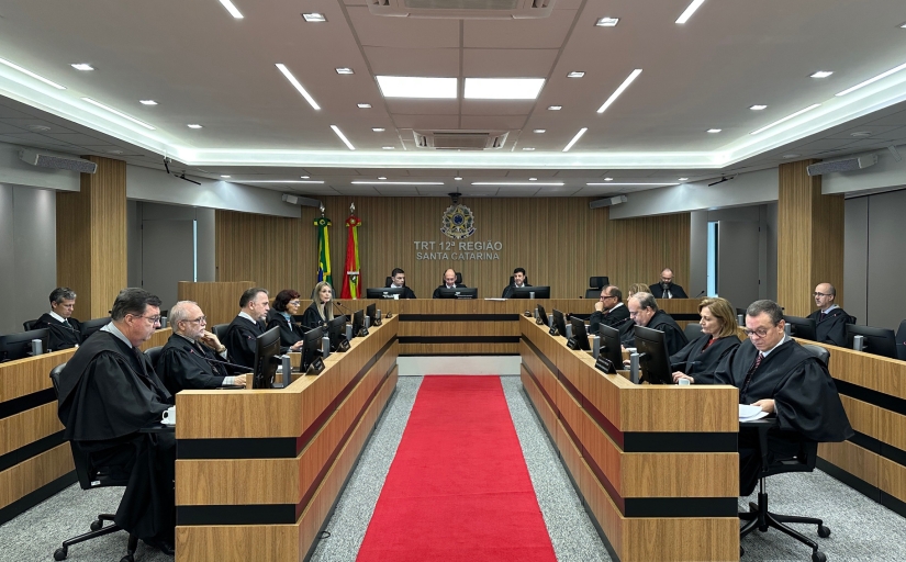 Fotografia em plano aberto da sala de sessão do Tribunal Pleno do TRT-SC. Ao centro, um corredor com um tapete na cor vermelha se estende até o fundo, onde há uma bancada onde está sentado o presidente da corte. Nas laterais direita e esquerda, há duas fileiras de postos de trabalho individuais, onde estão sentados os desembargadores.