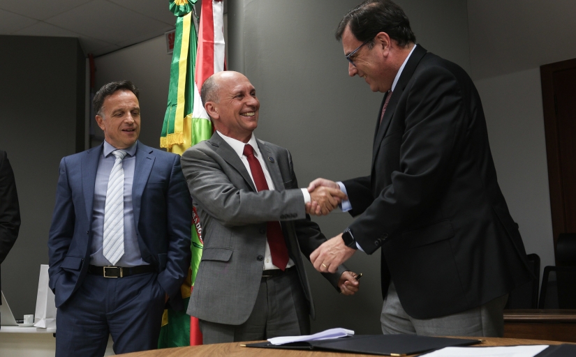 Dois homens de terno apertam as mãos sorrindo, em um ambiente formal com bandeiras ao fundo. Um terceiro homem ao lado, também de terno, observa a interação.