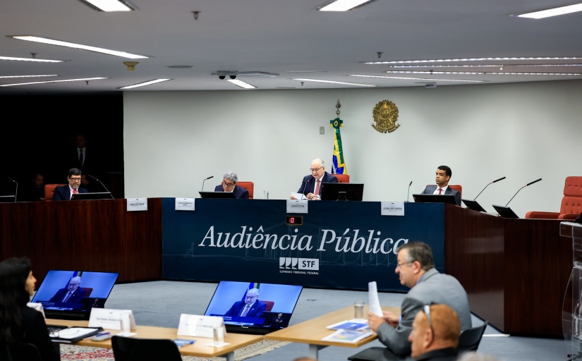 Plenário onde se encontram entre sete pessoas. Três homens se encontram ao centro da mesa na qual está escrito "audiência pública" e possui o logo do STF.