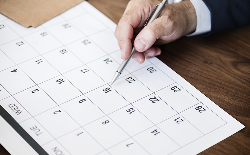 Fotografia em plano detalhe de uma mão segurando uma caneta apontando para uma data em um calendário de papel sobre uma mesa de madeira.