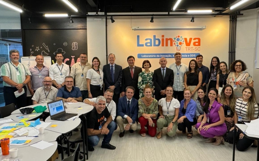 Grupo de 27 pessoas posando juntas em um espaço colaborativo com mesas, post-its e uma placa indicando "Labinova TRT-SC – Laboratório de Inovação".