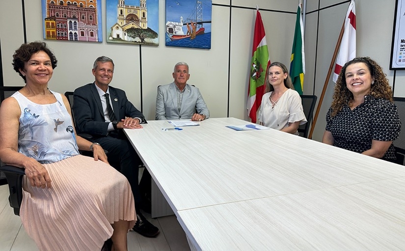 Cinco pessoas estão reunidas em uma sala, sentadas à mesa, com bandeiras e quadros ao fundo. 