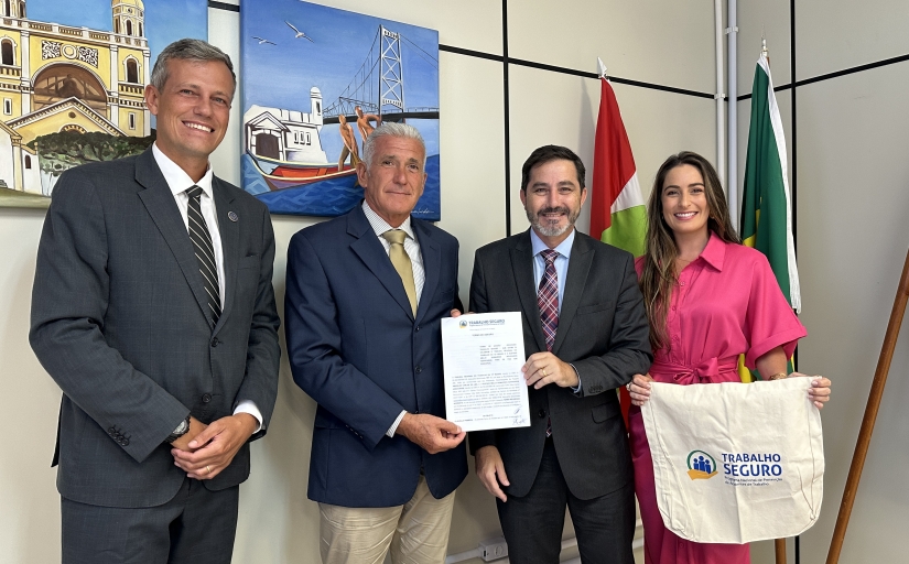 Quatro pessoas posando para foto, duas delas segurando um documento assinado e outra segurando uma bolsa com a logo do Programa "Trabalho Seguro". Ao fundo, três bandeiras