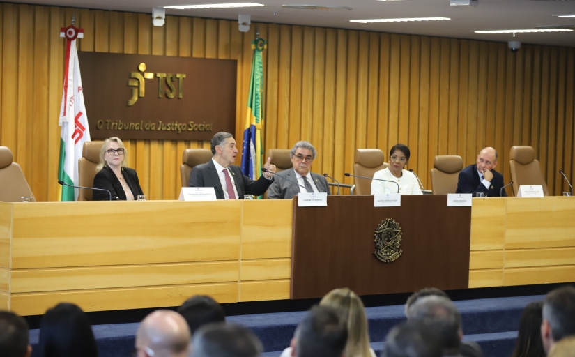 A imagem mostra uma mesa de autoridades no Tribunal Superior do Trabalho (TST), com placas nominais, bandeiras e um público acompanhando a sessão.