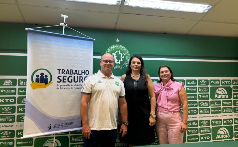 A imagem mostra um homem e duas mulheres pessoas posando para a foto em frente a um banner do Programa Trabalho Seguro,. O fundo é decorado com o escudo da Chapecoense e logomarcas de patrocinadores do clube.