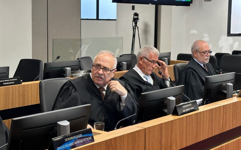 Fotografia da sessão em que houve a decisão. Três homens de toga estão sentados de frente para a tela de seus computadores em ambiente fechado.