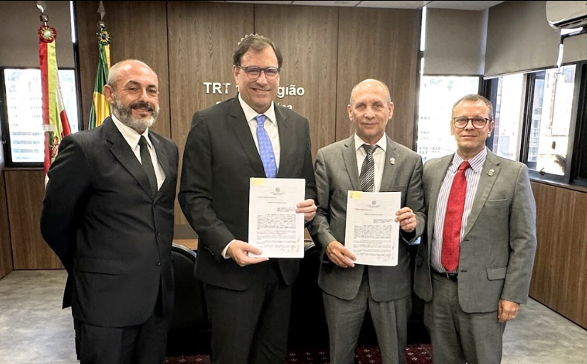 Quatro homens de terno e gravata estão em pé, posando e sorrindo para a foto, em uma sala de reuniões