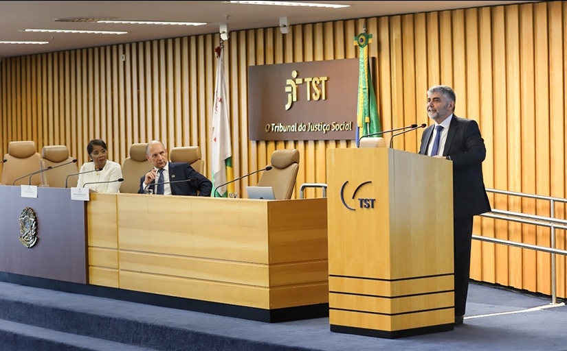 Homem de terno fala em um púlpito dentro de um auditório. Ao lado dele, um homem e uma mulher sentados em uma mesa o observam