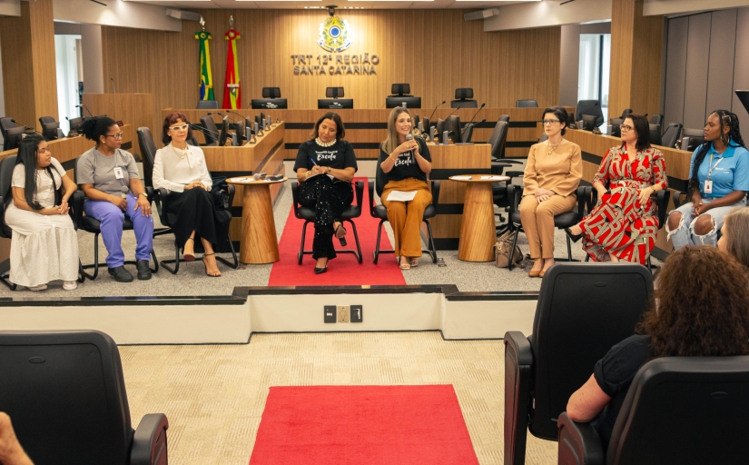 Oito mulheres sentadas em cadeiras, de frente para o público, em um evento formal