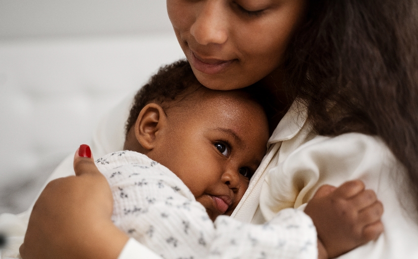 Fotografia ilustrativa de mulher negra com bebê no colo. Ambos vestem roupas claras.