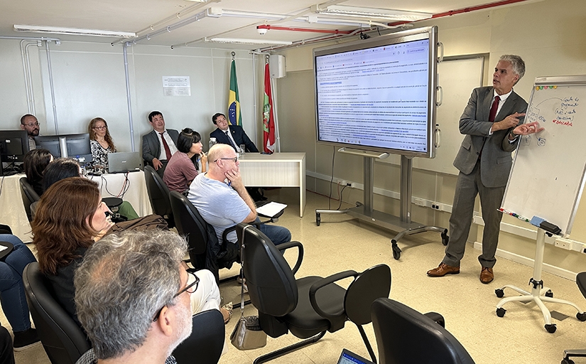 Em uma sala de aula, um homem branco de cabelos grisalhos, trajando um terno cinza, camisa branca e gravata vermelha, explica para um grupo de servidores, sentado em cadeiras, algo em um quadro branco. Na esquerda, há um monitor de vídeo, no qual consta o texto de uma lei.