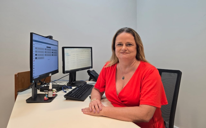 Uma mulher branca de cabelos loiros, usando uma blusa vermelha, está sentada atrás de uma mesa de cor clara, na qual há um computador com dois monitores e um telefone.