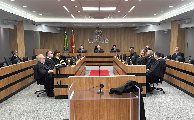 Em um plenário estão sentadas quinze pessoas, sendo três mulheres e doze homens. Eles vestem togas pretas e estão em estações de trabalho posicionadas em quatro fileiras. Ao fundo, estão sentadoes três homens, sendo ao centro, o presidente do TRT-SC.