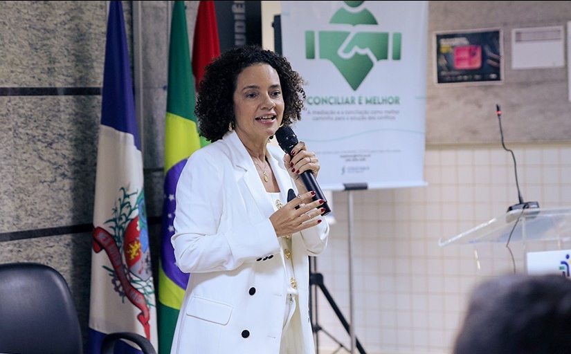 Uma mulher parda de cabelos escuros, curtos e cacheados, trajando um terno branco com calça e camisa brancas, fala em pé ao microfone em um auditório. Ao fundo, um banner onde se lê: Conciliar é Melhor.