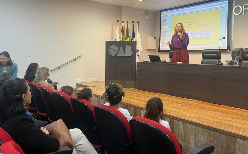 Mulher loira, de blusa rosa em pé diante de um auditório