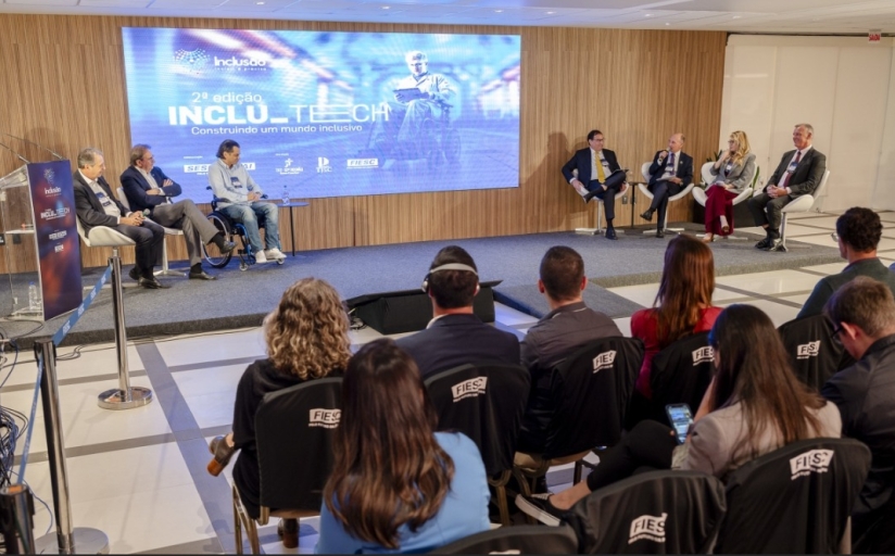 Seis homens e uma mulher estão sentados em um palco de um auditório dando uma palestra. Ao fundo um telão e na frente pessoas estão assistindo