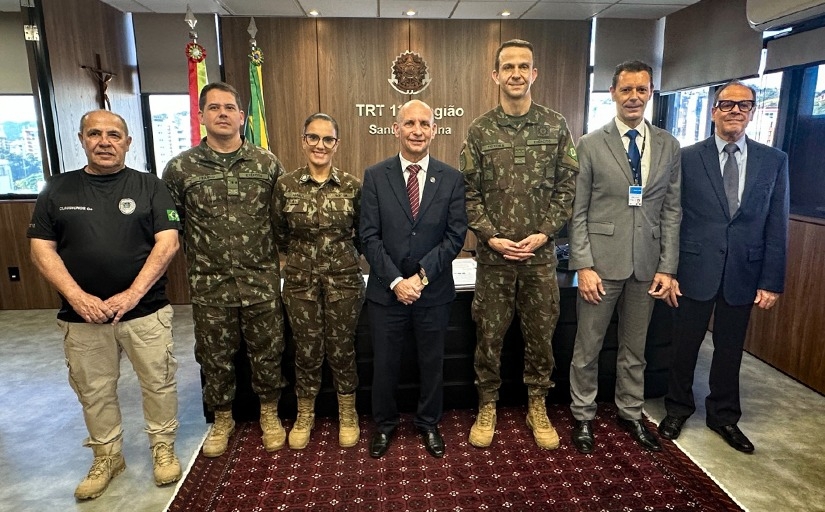 Sete pessoas em frente a uma escrivaninha posando para fotos. Três vestem terno e gravata e outros três uniformes do exército.